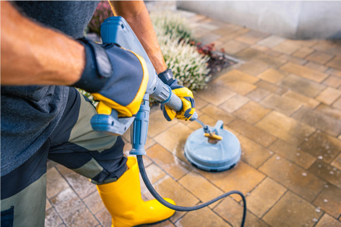Close up of a worker wearing yellow safety boots and protective gloves using a circular surface cleaner attachment to pressure wash stone patio pavers.