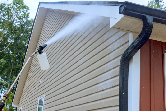 Pressure washer wand spraying cleaning detergent foam onto beige vinyl siding to safely soft wash the home exterior and remove mildew.